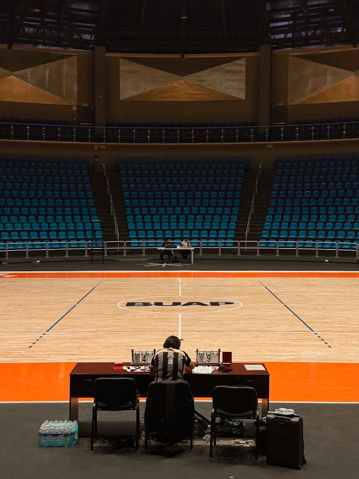 A lone official at a table on an empty basketball court with empty blue seats.